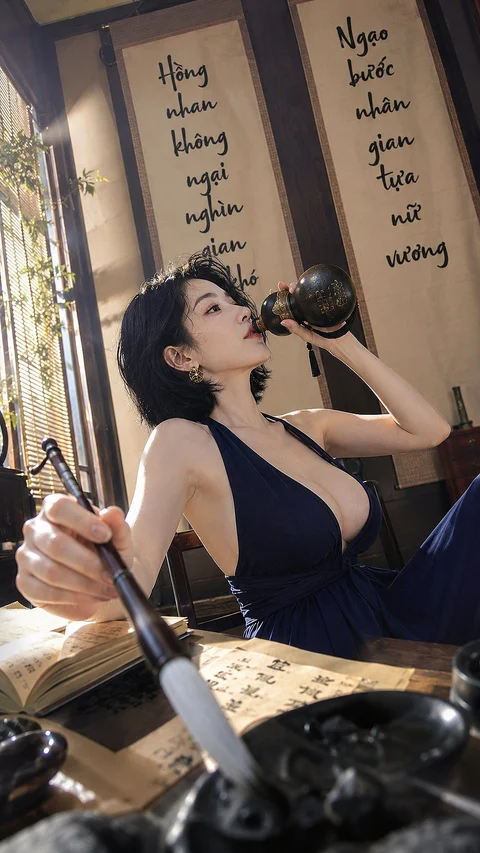 A woman in a dark-blue sleeveless outfit holds a large brush while drinking from a bottle beside hanging calligraphy.